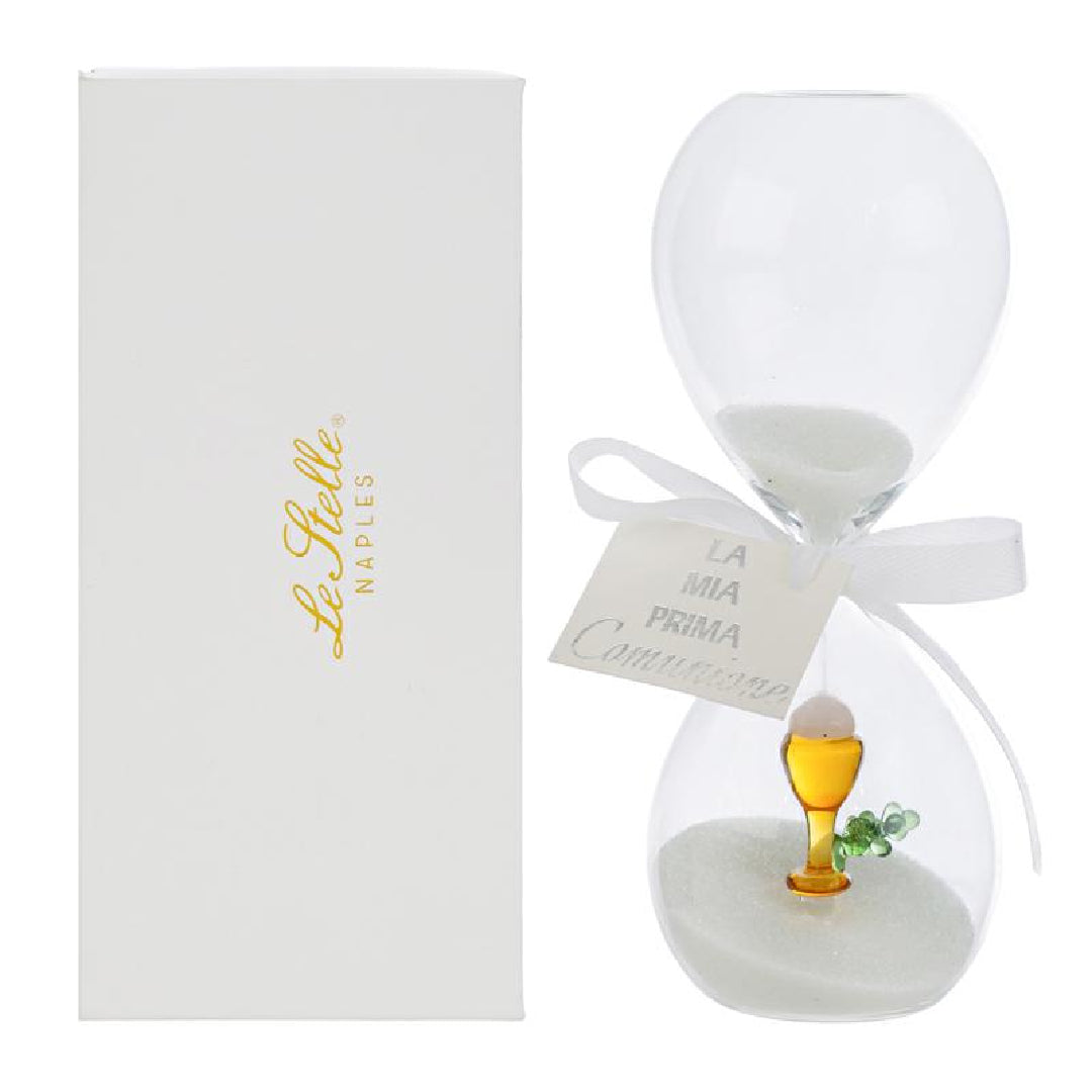 Clessidra in vetro trasparente riempita di sabbia bianca, con un calice dorato e un rametto verde all'interno del bulbo inferiore e fiocco bianco con etichetta "La mia prima Comunione". Bomboniera decorativa presentata con la sua confezione bianca del marchio Le Stelle, ideale come ricordo per la Prima Comunione.