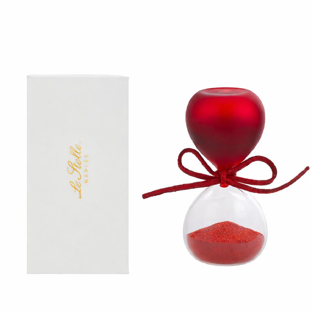 Clessidra in vetro satinato rosso a forma di clava, con base conica trasparente riempita di sabbia rossa e cordoncino con etichetta tonda "Le Stelle" decorata da un cappello di laurea. Oggetto decorativo elegante presentato con la sua confezione bianca del marchio, ideale come bomboniera per lauree o idea regalo raffinata.