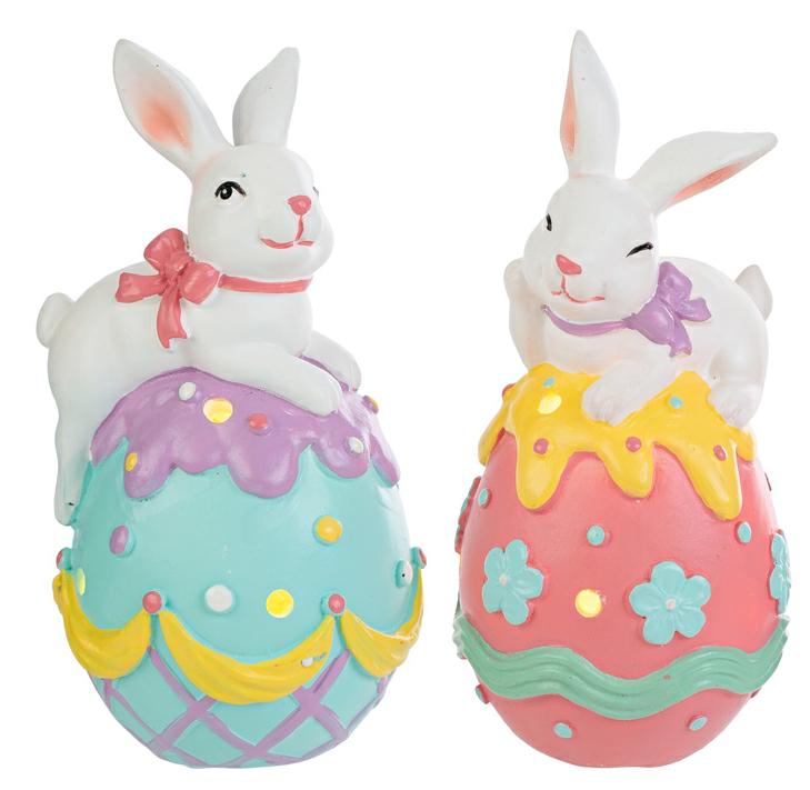 Coppia di statuine pasquali in resina con LED. Ogni soggetto raffigura un coniglietto bianco disteso sopra un grande uovo decorato con colori vivaci, fiori e motivi a rilievo. Le uova presentano piccoli fori circolari da cui filtra la luce interna. Una versione ha l'uovo azzurro e viola, l'altra rosa e giallo.