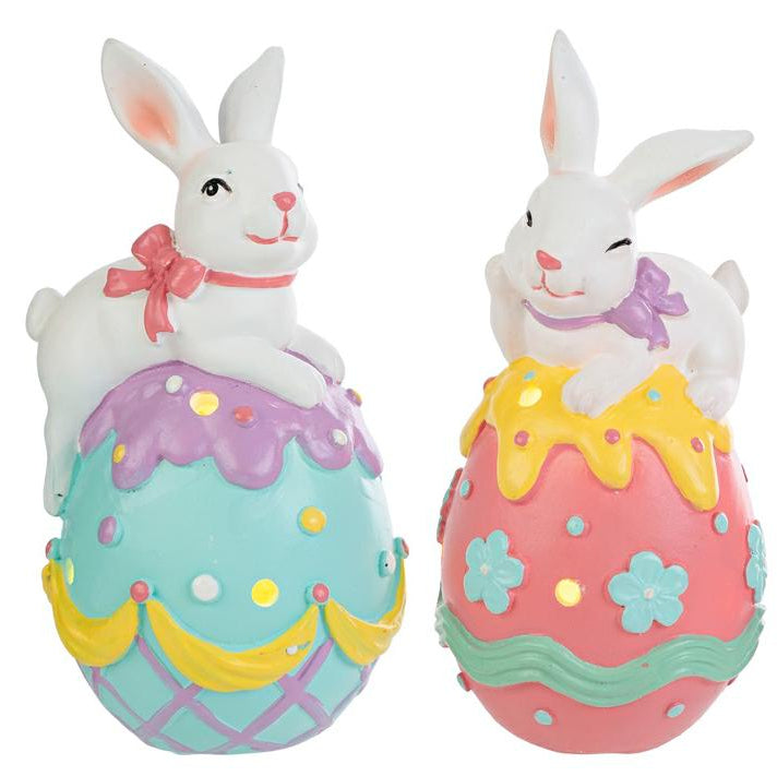 Coppia di statuine pasquali in resina con LED. Ogni soggetto raffigura un coniglietto bianco disteso sopra un grande uovo decorato con colori vivaci, fiori e motivi a rilievo. Le uova presentano piccoli fori circolari da cui filtra la luce interna. Una versione ha l'uovo azzurro e viola, l'altra rosa e giallo.