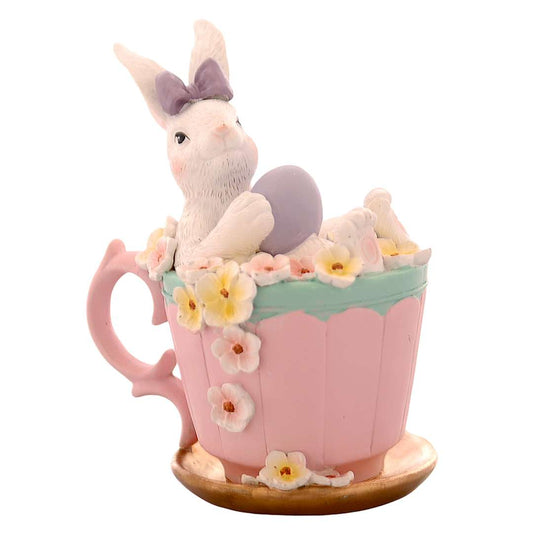 Decorazione pasquale Vetur in poliresina. Coniglietto bianco con fiocco viola seduto in una tazza rosa decorata con fiori gialli e bianchi, mentre regge un uovo viola. Altezza 16,5 cm.