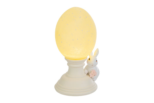 Lampada decorativa pasquale a forma di uovo luminoso giallo crema con effetto puntinato su piedistallo bianco. Accanto alla base, una statuina di coniglietto bianco che stringe un piccolo mazzo di fiori rosa. Oggetto d'arredo per Pasqua 2026 del marchio Le Stelle, ideale come luce notturna o decorazione da tavolo.