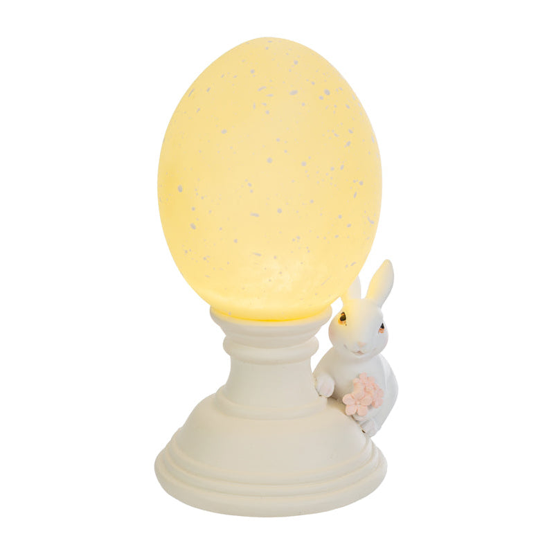 Lampada decorativa pasquale a forma di uovo luminoso giallo crema con effetto puntinato su piedistallo bianco. Accanto alla base, una statuina di coniglietto bianco che stringe un piccolo mazzo di fiori rosa. Oggetto d'arredo per Pasqua 2026 del marchio Le Stelle, ideale come luce notturna o decorazione da tavolo.