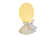 Lampada decorativa pasquale a forma di uovo luminoso giallo crema con effetto puntinato su piedistallo bianco. Accanto alla base, una statuina di coniglietto bianco che stringe un piccolo mazzo di fiori rosa. Oggetto d'arredo per Pasqua 2026 del marchio Le Stelle, ideale come luce notturna o decorazione da tavolo.