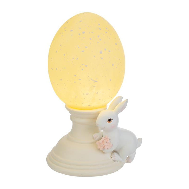 Lampada decorativa pasquale a forma di uovo luminoso giallo crema con effetto puntinato su piedistallo bianco. Accanto alla base, una statuina di coniglietto bianco che stringe un piccolo mazzo di fiori rosa. Oggetto d'arredo per Pasqua 2026 del marchio Le Stelle, ideale come luce notturna o decorazione da tavolo.