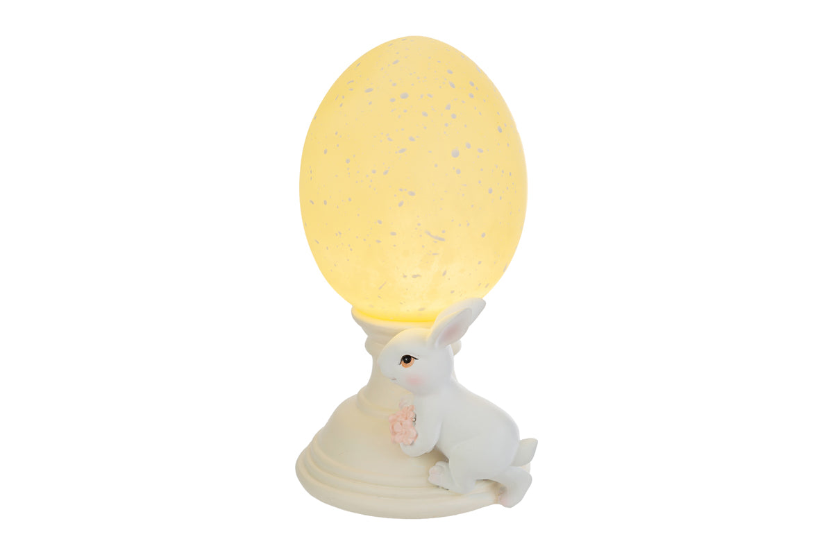 Lampada decorativa pasquale a forma di uovo luminoso giallo crema con effetto puntinato su piedistallo bianco. Accanto alla base, una statuina di coniglietto bianco che stringe un piccolo mazzo di fiori rosa. Oggetto d'arredo per Pasqua 2026 del marchio Le Stelle, ideale come luce notturna o decorazione da tavolo.