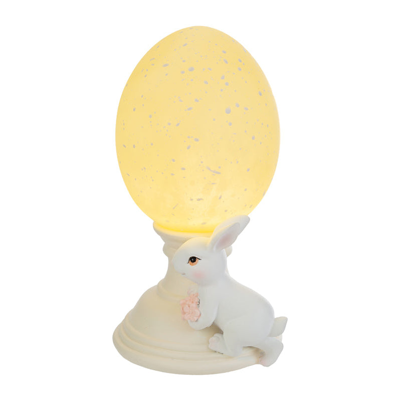 Lampada decorativa pasquale a forma di uovo luminoso giallo crema con effetto puntinato su piedistallo bianco. Accanto alla base, una statuina di coniglietto bianco che stringe un piccolo mazzo di fiori rosa. Oggetto d'arredo per Pasqua 2026 del marchio Le Stelle, ideale come luce notturna o decorazione da tavolo.