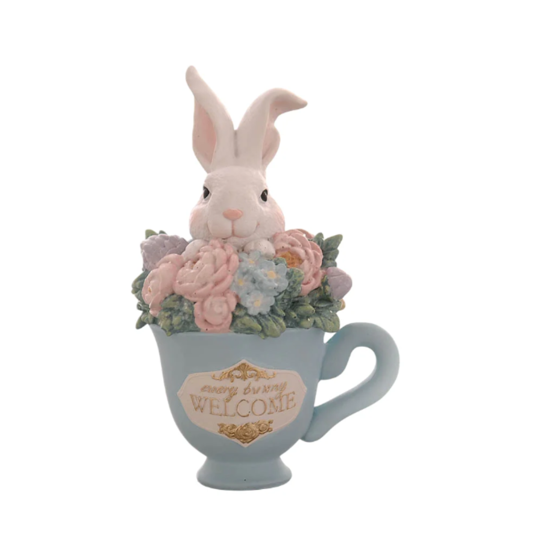 Statuina decorativa in resina raffigurante un coniglietto bianco che spunta da una tazzina azzurra ricolma di fiori primaverili (rose rosa e piccoli fiori azzurri). La tazzina presenta una placca frontale bianca con bordi dorati e la scritta "Every bunny Welcome". Design curato con dettagli floreali e colori pastello.