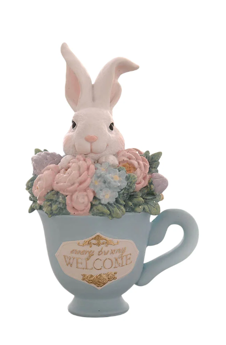 Statuina decorativa in resina raffigurante un coniglietto bianco che spunta da una tazzina azzurra ricolma di fiori primaverili (rose rosa e piccoli fiori azzurri). La tazzina presenta una placca frontale bianca con bordi dorati e la scritta "Every bunny Welcome". Design curato con dettagli floreali e colori pastello.