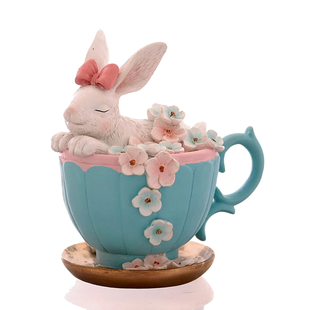 Decorazione pasquale scultorea Vetur in poliresina. Coniglietto bianco con fiocco rosaseduto all'interno di una tazza da tè azzurra decorata con fiori rosa e celesti sfumati, appoggiata su un piattino dorato.