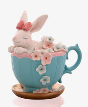 Decorazione pasquale scultorea Vetur in poliresina. Coniglietto bianco con fiocco rosaseduto all'interno di una tazza da tè azzurra decorata con fiori rosa e celesti sfumati, appoggiata su un piattino dorato.