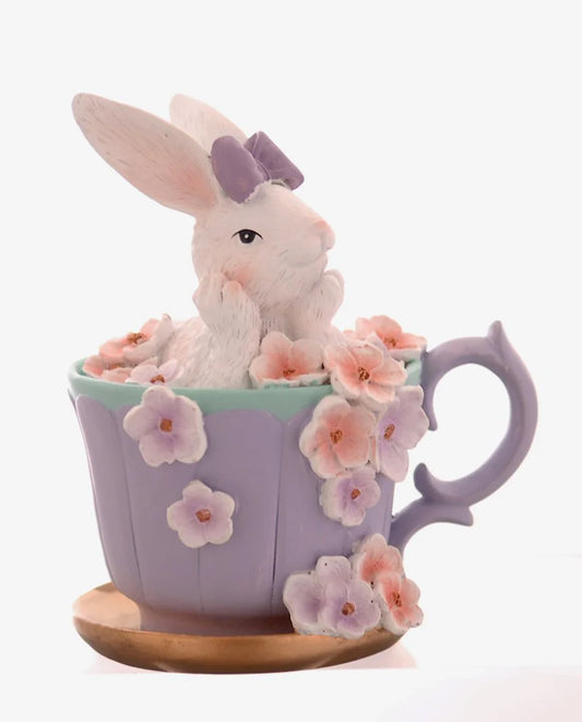 Decorazione pasquale scultorea Vetur in poliresina. Coniglietto bianco con fiocco viola seduto all'interno di una tazza da tè lilla decorata con fiori rosa e viola sfumati, appoggiata su un piattino dorato.