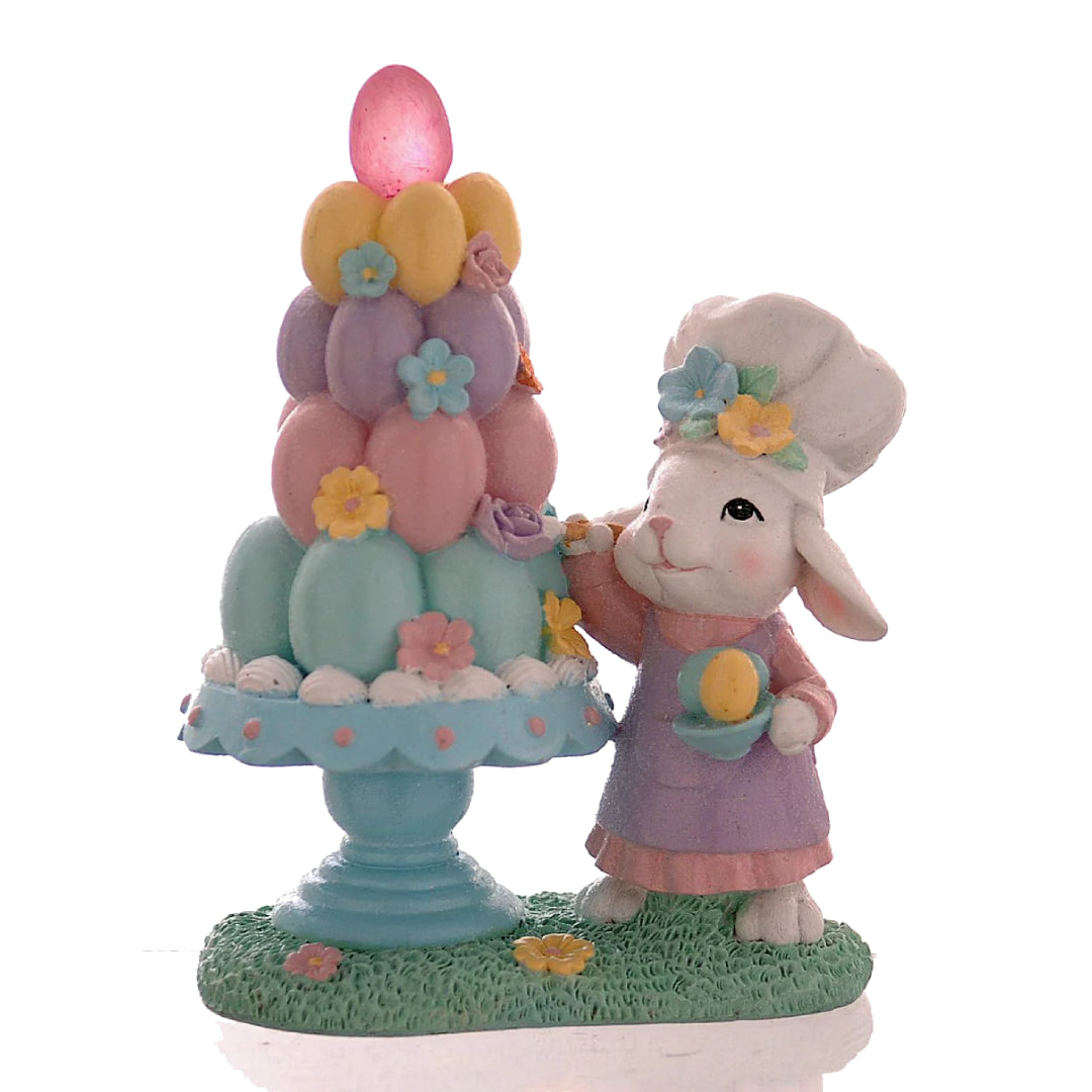 Decorazione pasquale Vetur in poliresina h 21 cm. Scenetta con coniglio bianco pasticcere che decora una piramide di uova colorate su un'alzatina azzurra, dotata di luce LED interna.