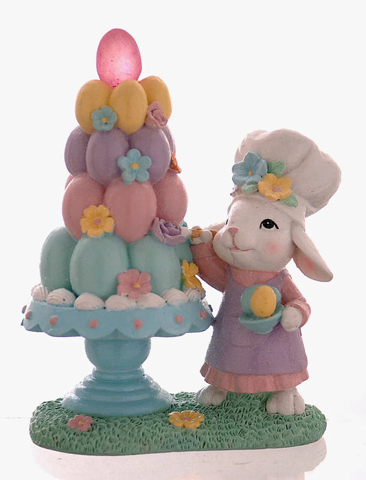 Decorazione pasquale Vetur in poliresina h 21 cm. Scenetta con coniglio bianco pasticcere che decora una piramide di uova colorate su un'alzatina azzurra, dotata di luce LED interna.