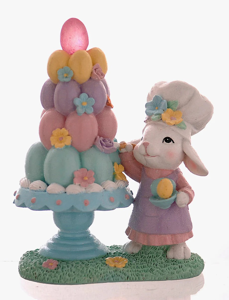 Decorazione pasquale Vetur in poliresina h 21 cm. Scenetta con coniglio bianco pasticcere che decora una piramide di uova colorate su un'alzatina azzurra, dotata di luce LED interna.
