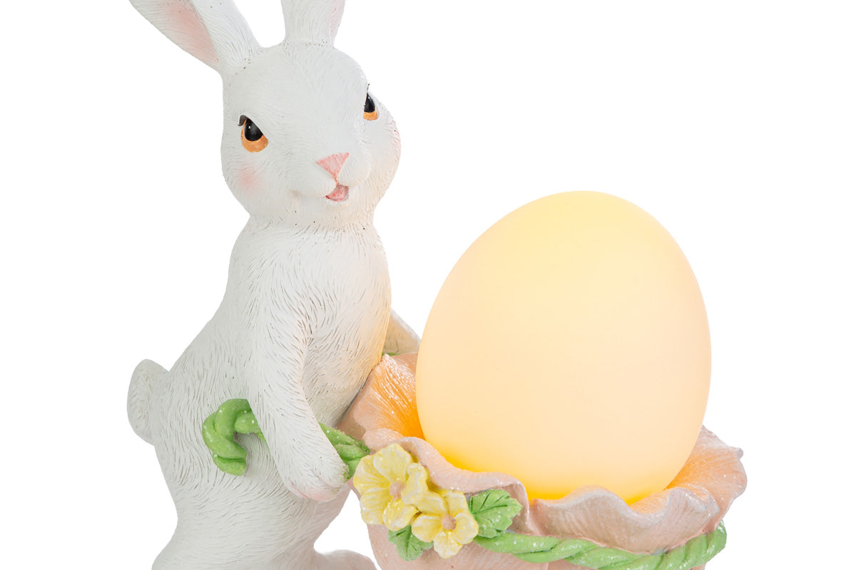 Statuina pasquale luminosa del marchio Le Stelle con un coniglietto bianco in piedi che spinge una carriola a forma di guscio d'uovo rosa. La carriola, decorata con fiorellini gialli, contiene un uovo luminoso giallo acceso. Decorazione in resina per Pasqua 2026 con dettagli realistici del pelo e del volto.