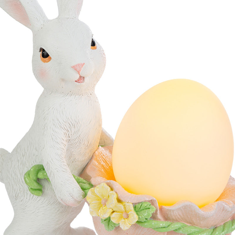 Statuina pasquale luminosa del marchio Le Stelle con un coniglietto bianco in piedi che spinge una carriola a forma di guscio d'uovo rosa. La carriola, decorata con fiorellini gialli, contiene un uovo luminoso giallo acceso. Decorazione in resina per Pasqua 2026 con dettagli realistici del pelo e del volto.
