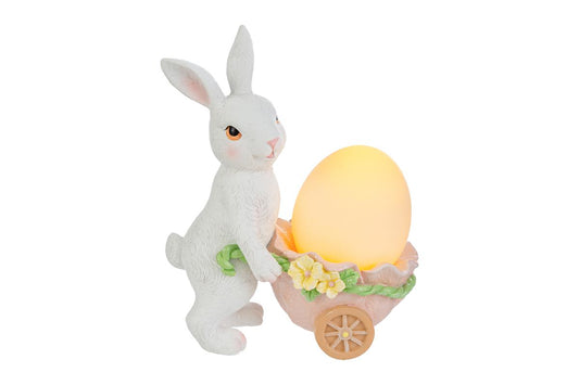 Statuina pasquale luminosa del marchio Le Stelle con un coniglietto bianco in piedi che spinge una carriola a forma di guscio d'uovo rosa. La carriola, decorata con fiorellini gialli, contiene un uovo luminoso giallo acceso. Decorazione in resina per Pasqua 2026 con dettagli realistici del pelo e del volto.