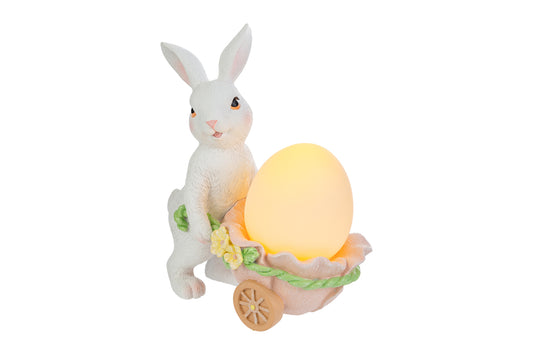 Statuina pasquale luminosa del marchio Le Stelle con un coniglietto bianco in piedi che spinge una carriola a forma di guscio d'uovo rosa. La carriola, decorata con fiorellini gialli, contiene un uovo luminoso giallo acceso. Decorazione in resina per Pasqua 2026 con dettagli realistici del pelo e del volto.