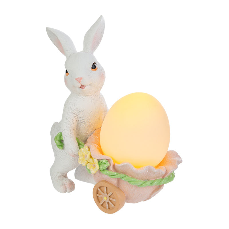 Statuina pasquale luminosa del marchio Le Stelle con un coniglietto bianco in piedi che spinge una carriola a forma di guscio d'uovo rosa. La carriola, decorata con fiorellini gialli, contiene un uovo luminoso giallo acceso. Decorazione in resina per Pasqua 2026 con dettagli realistici del pelo e del volto.