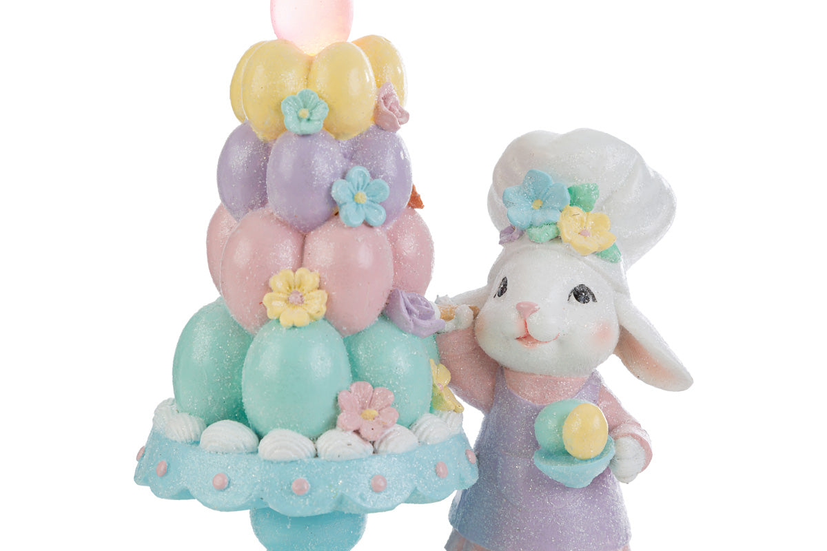 Statuina decorativa pasquale del marchio "Le Stelle" raffigurante un coniglio chef accanto a un albero di Pasqua stilizzato con uova colorate dotate di luci LED. Il coniglio indossa un cappello da cuoco e una giacca rosa, mentre l'albero presenta uova decorate in tonalità pastello giallo, rosa e azzurro su una base di prato verde.
