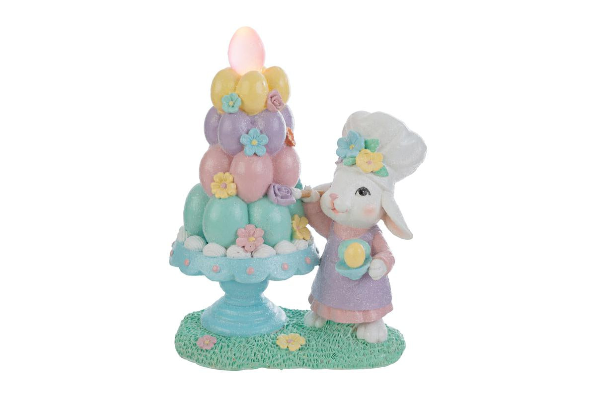 Statuina decorativa pasquale del marchio "Le Stelle" raffigurante un coniglio chef accanto a un albero di Pasqua stilizzato con uova colorate dotate di luci LED. Il coniglio indossa un cappello da cuoco e una giacca rosa, mentre l'albero presenta uova decorate in tonalità pastello giallo, rosa e azzurro su una base di prato verde.