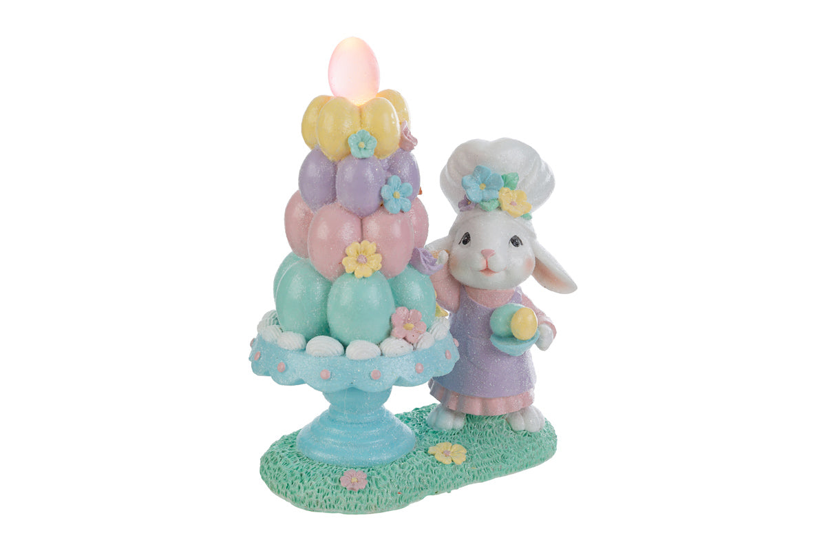 Statuina decorativa pasquale del marchio "Le Stelle" raffigurante un coniglio chef accanto a un albero di Pasqua stilizzato con uova colorate dotate di luci LED. Il coniglio indossa un cappello da cuoco e una giacca rosa, mentre l'albero presenta uova decorate in tonalità pastello giallo, rosa e azzurro su una base di prato verde.
