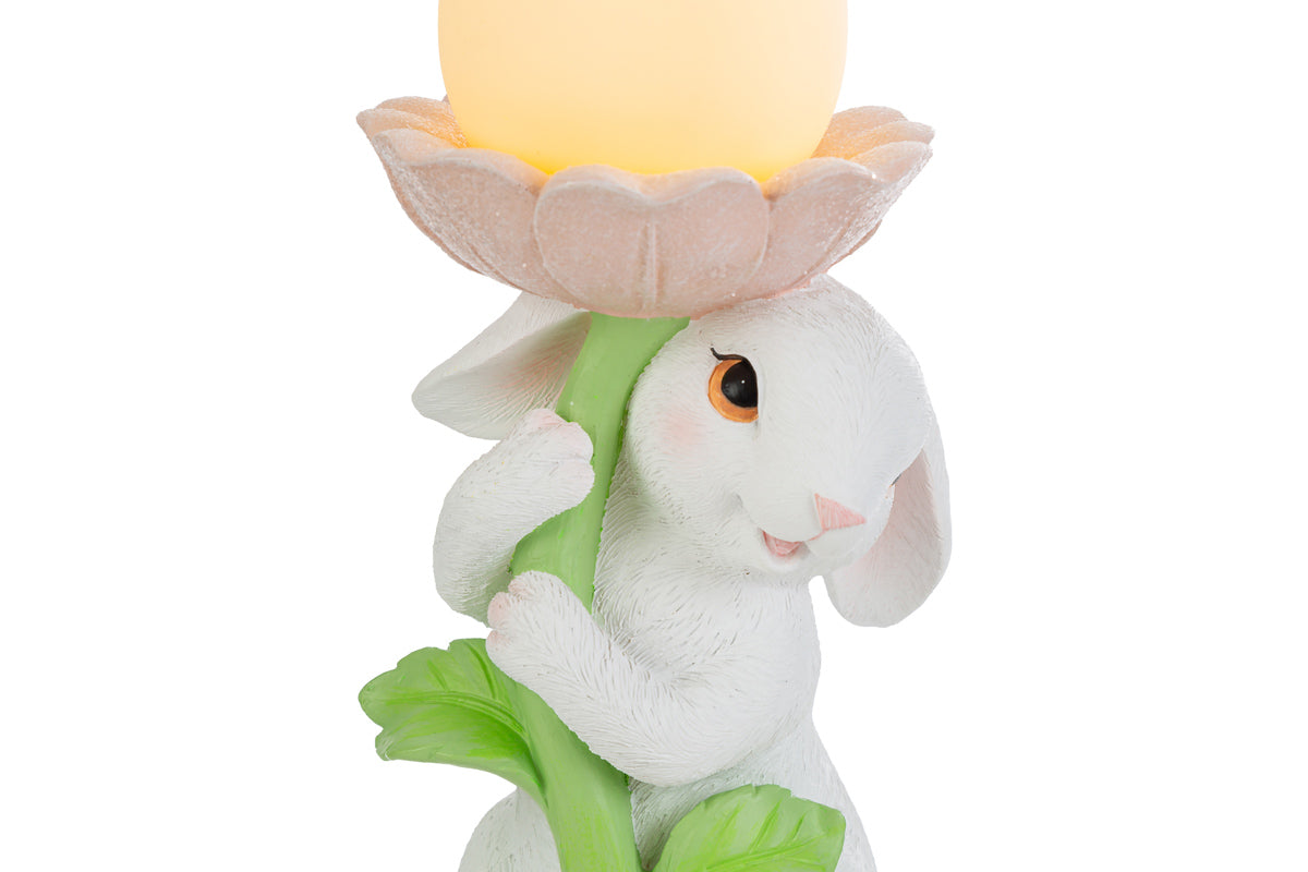 Statuina pasquale del marchio Le Stelle raffigurante un coniglietto bianco che abbraccia lo stelo verde di un grande fiore rosa. All'interno della corolla del fiore è posizionato un uovo luminoso a LED giallo che funge da lampada. Decorazione in resina per Pasqua 2026 con finitura dettagliata e colori pastello.