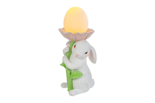 Statuina pasquale del marchio Le Stelle raffigurante un coniglietto bianco che abbraccia lo stelo verde di un grande fiore rosa. All'interno della corolla del fiore è posizionato un uovo luminoso a LED giallo che funge da lampada. Decorazione in resina per Pasqua 2026 con finitura dettagliata e colori pastello.