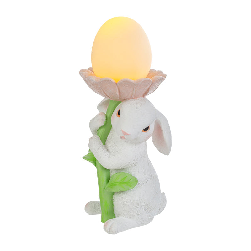 Statuina pasquale del marchio Le Stelle raffigurante un coniglietto bianco che abbraccia lo stelo verde di un grande fiore rosa. All'interno della corolla del fiore è posizionato un uovo luminoso a LED giallo che funge da lampada. Decorazione in resina per Pasqua 2026 con finitura dettagliata e colori pastello.
