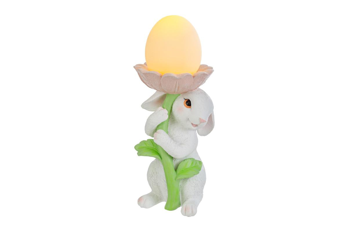 Statuina pasquale del marchio Le Stelle raffigurante un coniglietto bianco che abbraccia lo stelo verde di un grande fiore rosa. All'interno della corolla del fiore è posizionato un uovo luminoso a LED giallo che funge da lampada. Decorazione in resina per Pasqua 2026 con finitura dettagliata e colori pastello.