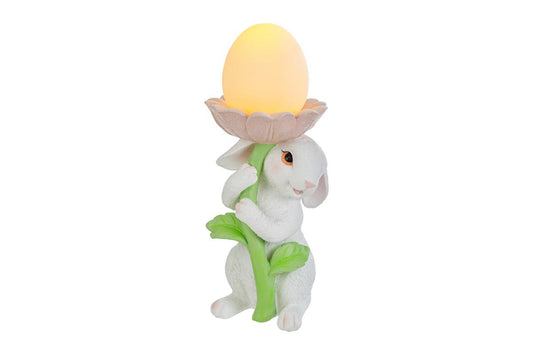 Statuina pasquale del marchio Le Stelle raffigurante un coniglietto bianco che abbraccia lo stelo verde di un grande fiore rosa. All'interno della corolla del fiore è posizionato un uovo luminoso a LED giallo che funge da lampada. Decorazione in resina per Pasqua 2026 con finitura dettagliata e colori pastello.