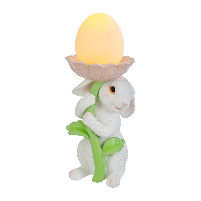 Statuina pasquale del marchio Le Stelle raffigurante un coniglietto bianco che abbraccia lo stelo verde di un grande fiore rosa. All'interno della corolla del fiore è posizionato un uovo luminoso a LED giallo che funge da lampada. Decorazione in resina per Pasqua 2026 con finitura dettagliata e colori pastello.