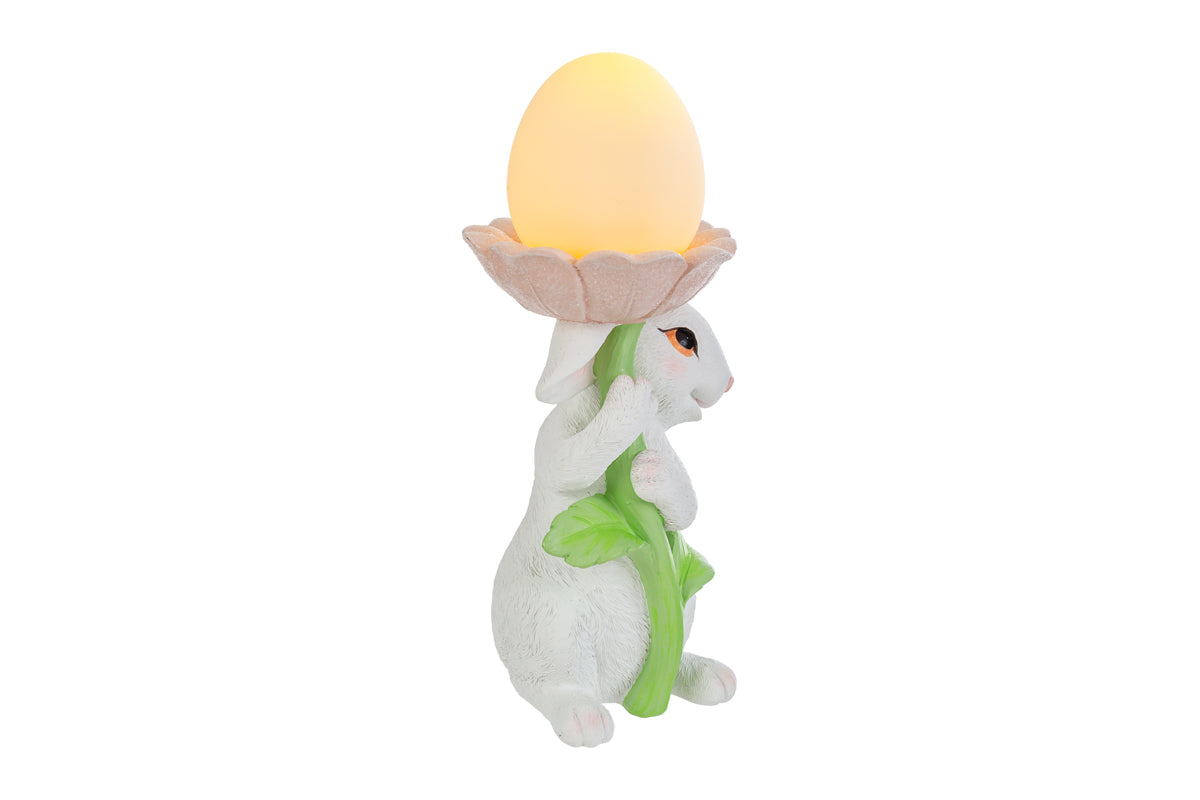 Statuina pasquale del marchio Le Stelle raffigurante un coniglietto bianco che abbraccia lo stelo verde di un grande fiore rosa. All'interno della corolla del fiore è posizionato un uovo luminoso a LED giallo che funge da lampada. Decorazione in resina per Pasqua 2026 con finitura dettagliata e colori pastello.