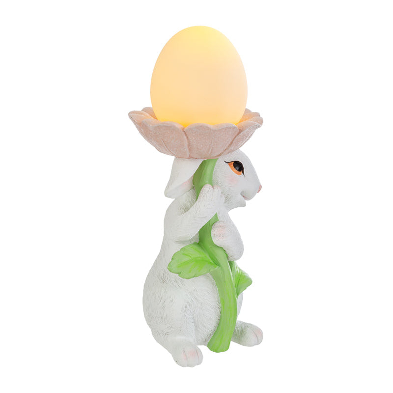 Statuina pasquale del marchio Le Stelle raffigurante un coniglietto bianco che abbraccia lo stelo verde di un grande fiore rosa. All'interno della corolla del fiore è posizionato un uovo luminoso a LED giallo che funge da lampada. Decorazione in resina per Pasqua 2026 con finitura dettagliata e colori pastello.