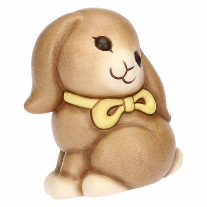 Statuina in ceramica Thun raffigurante il coniglio Roger della collezione Incanto di Pasqua. Il coniglietto, nei classici toni del marrone sfumato, è ritratto seduto con un fiocco giallo al collo e grandi occhi dolci. Iconica decorazione pasquale Thun con finitura opaca e forme morbide.