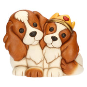 Statuina in ceramica Thun raffigurante una coppia di cani Cavalier King accovacciati uno accanto all'altro. Sono dipinti nei toni del bianco e del marrone caldo; il cane a destra indossa una corona dorata decorata con un piccolo cuore rosso. Lo stile è quello classico e tondeggiante tipico del brand.