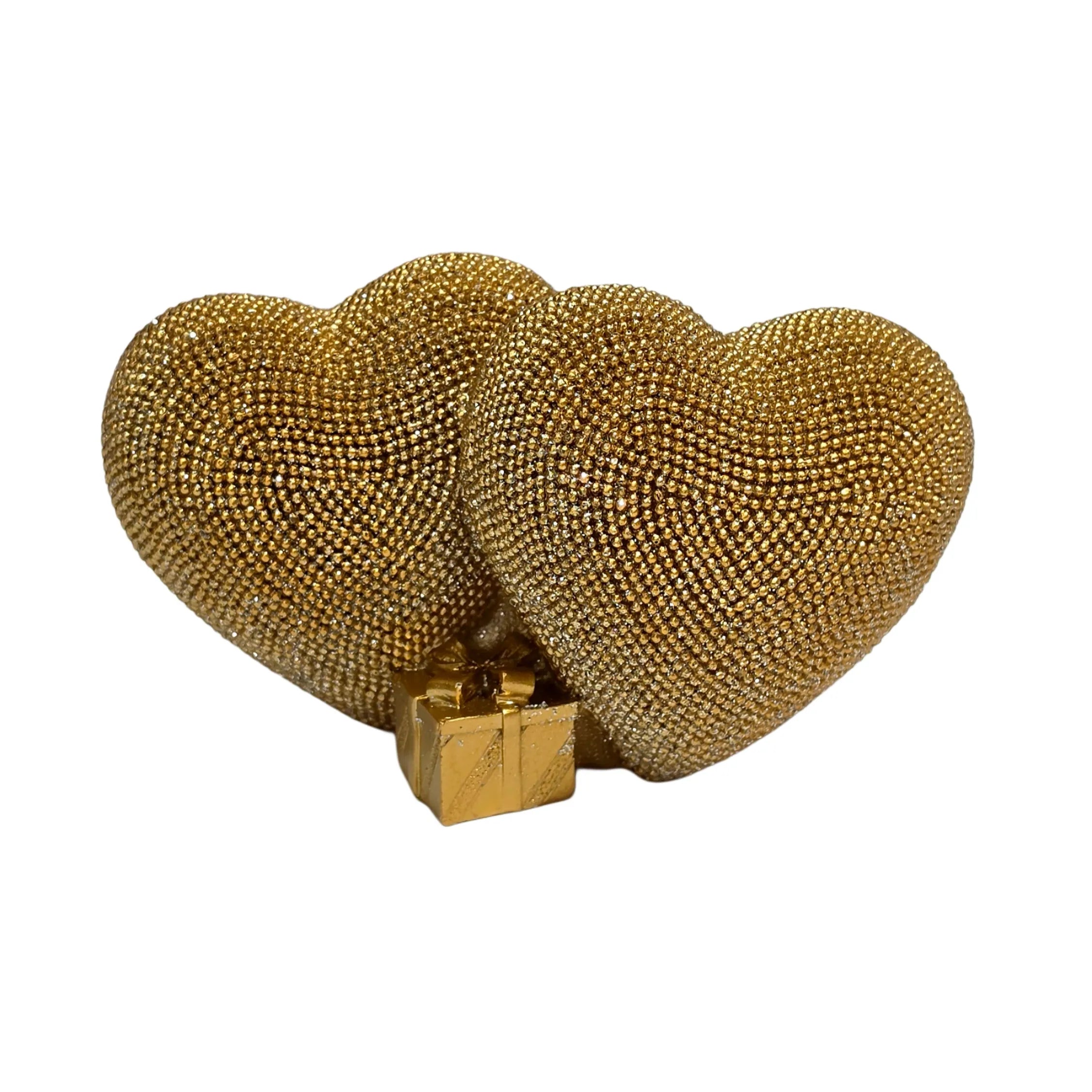 Statuina in resina raffigurante due cuori dorati affiancati, interamente ricoperti di brillantini glitterati, con un piccolo pacco regalo dorato liscio incastonato alla base. Oggetto decorativo firmato Sud Import, ideale come soprammobile romantico o idea regalo per San Valentino e occasioni speciali.