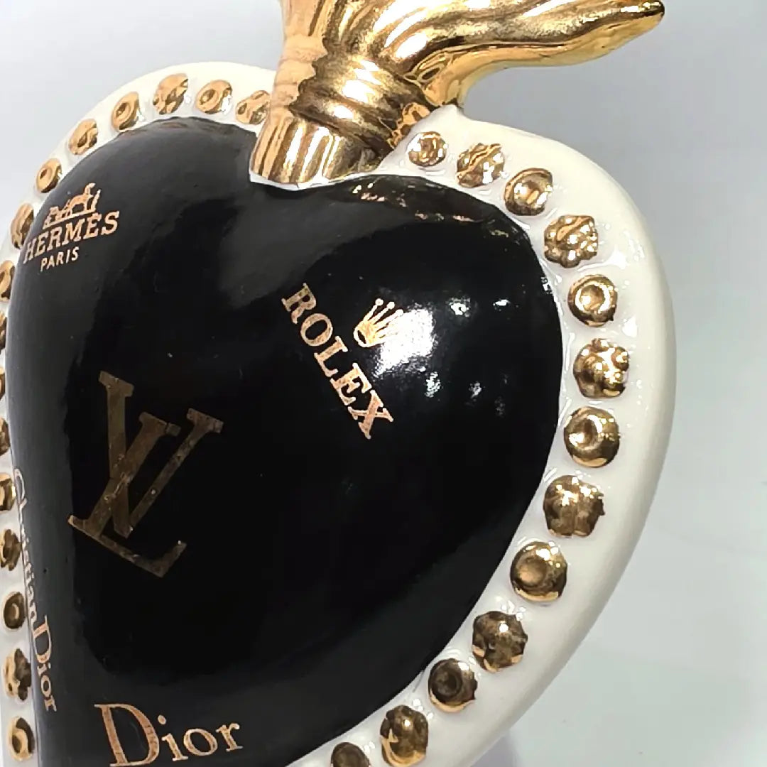 Scultura in ceramica a forma di Cuore Sacro firmata Sbordone, caratterizzata da un corpo nero lucido decorato con loghi fashion dorati e una fiamma superiore in oro. L'opera è rifinita da una cornice bianca perlinata con borchie dorate, rappresentando un complemento d'arredo pop art esclusivo e moderno per interni di design.