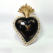 Scultura in ceramica a forma di Cuore Sacro firmata Sbordone, caratterizzata da un corpo nero lucido decorato con loghi fashion dorati e una fiamma superiore in oro. L'opera è rifinita da una cornice bianca perlinata con borchie dorate, rappresentando un complemento d'arredo pop art esclusivo e moderno per interni di design.