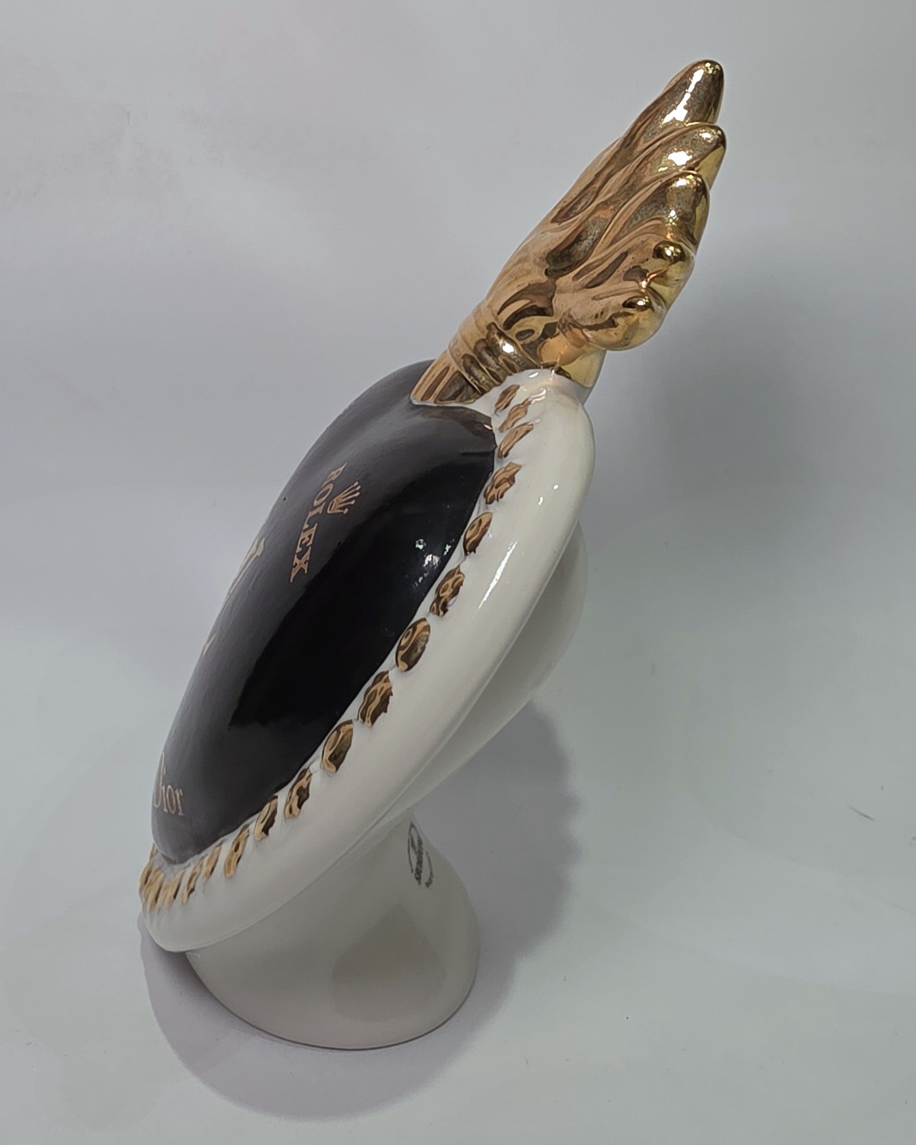 Scultura in ceramica a forma di Cuore Sacro firmata Sbordone, caratterizzata da un corpo nero lucido decorato con loghi fashion dorati e una fiamma superiore in oro. L'opera è rifinita da una cornice bianca perlinata con borchie dorate, rappresentando un complemento d'arredo pop art esclusivo e moderno per interni di design.