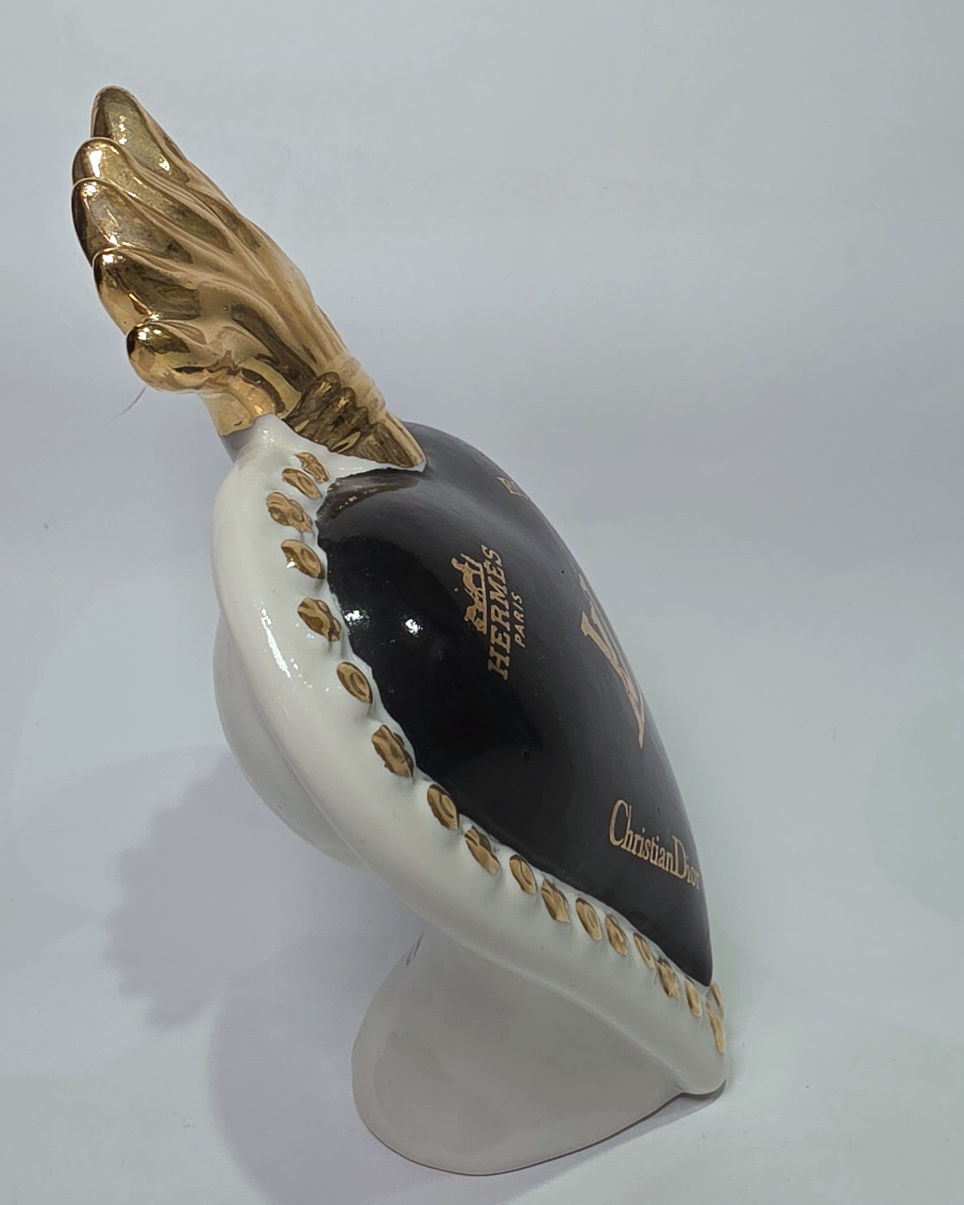 Scultura in ceramica a forma di Cuore Sacro firmata Sbordone, caratterizzata da un corpo nero lucido decorato con loghi fashion dorati e una fiamma superiore in oro. L'opera è rifinita da una cornice bianca perlinata con borchie dorate, rappresentando un complemento d'arredo pop art esclusivo e moderno per interni di design.