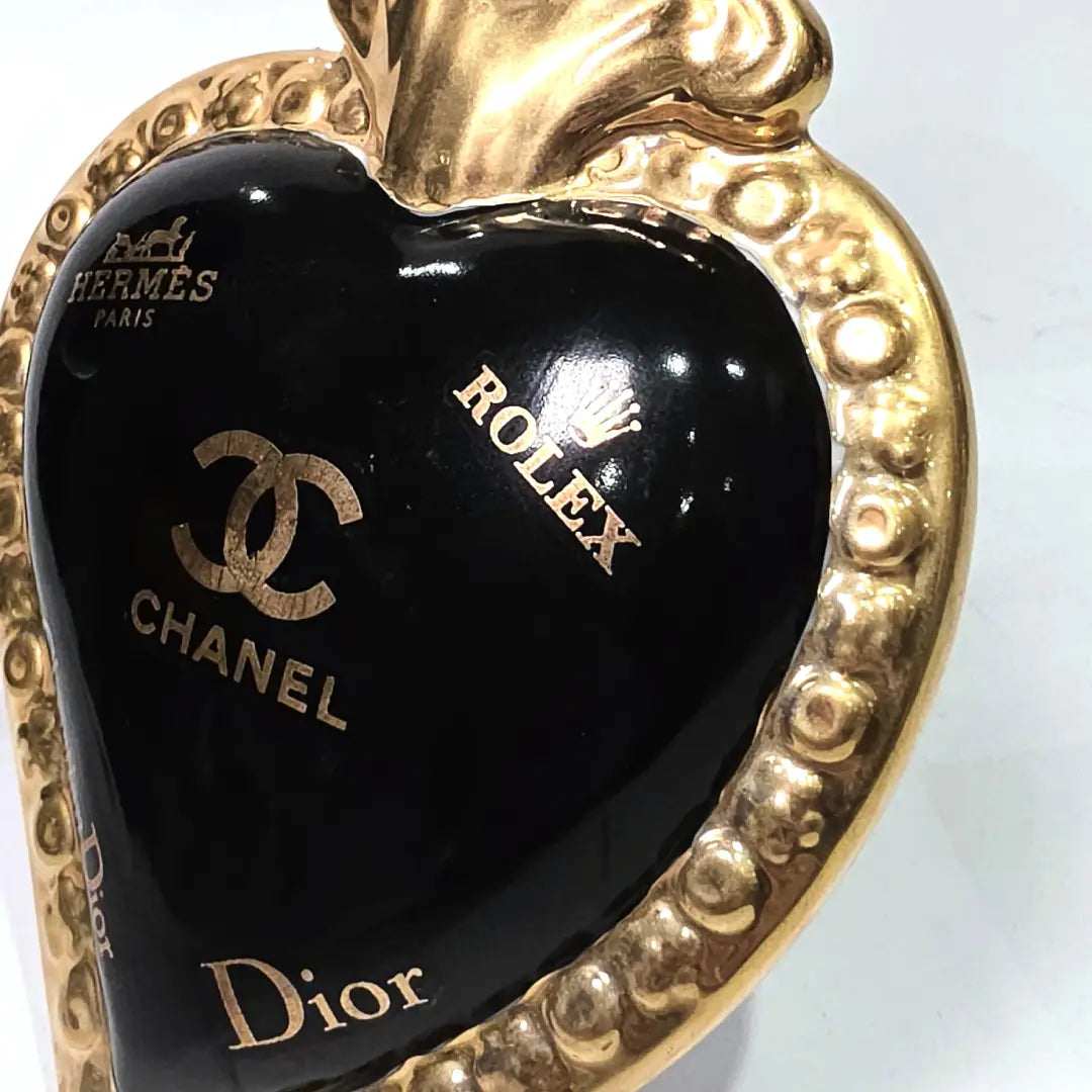 Scultura a forma di Cuore Sacro in ceramica nera lucida, impreziosita da una ricca cornice dorata lavorata a sbalzo e decorata con loghi fashion iconici in oro. Questo esclusivo oggetto d'arredo in stile pop art presenta una fiamma superiore dorata e una base d'appoggio integrata, perfetto per aggiungere un dettaglio di lusso moderno agli ambienti interni.