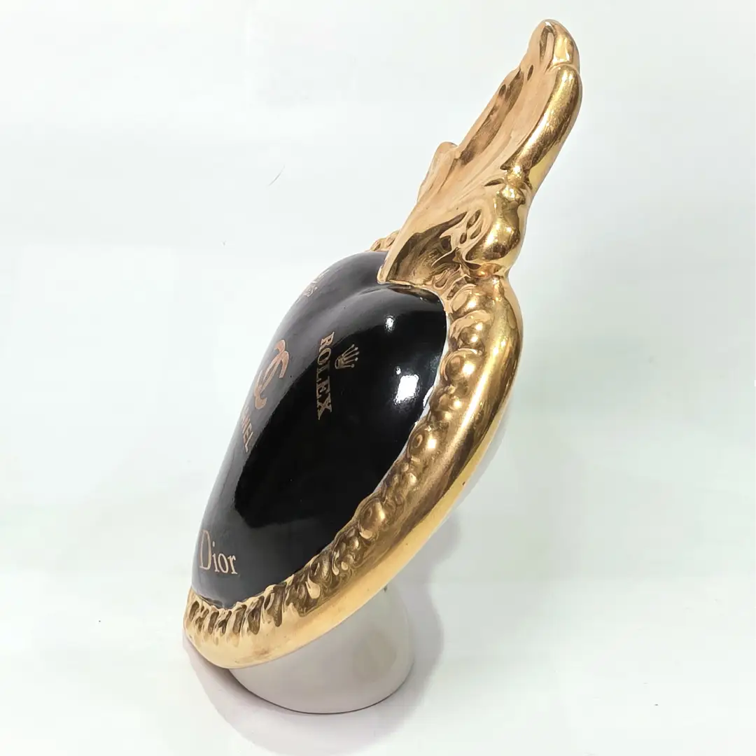 Scultura a forma di Cuore Sacro in ceramica nera lucida, impreziosita da una ricca cornice dorata lavorata a sbalzo e decorata con loghi fashion iconici in oro. Questo esclusivo oggetto d'arredo in stile pop art presenta una fiamma superiore dorata e una base d'appoggio integrata, perfetto per aggiungere un dettaglio di lusso moderno agli ambienti interni.