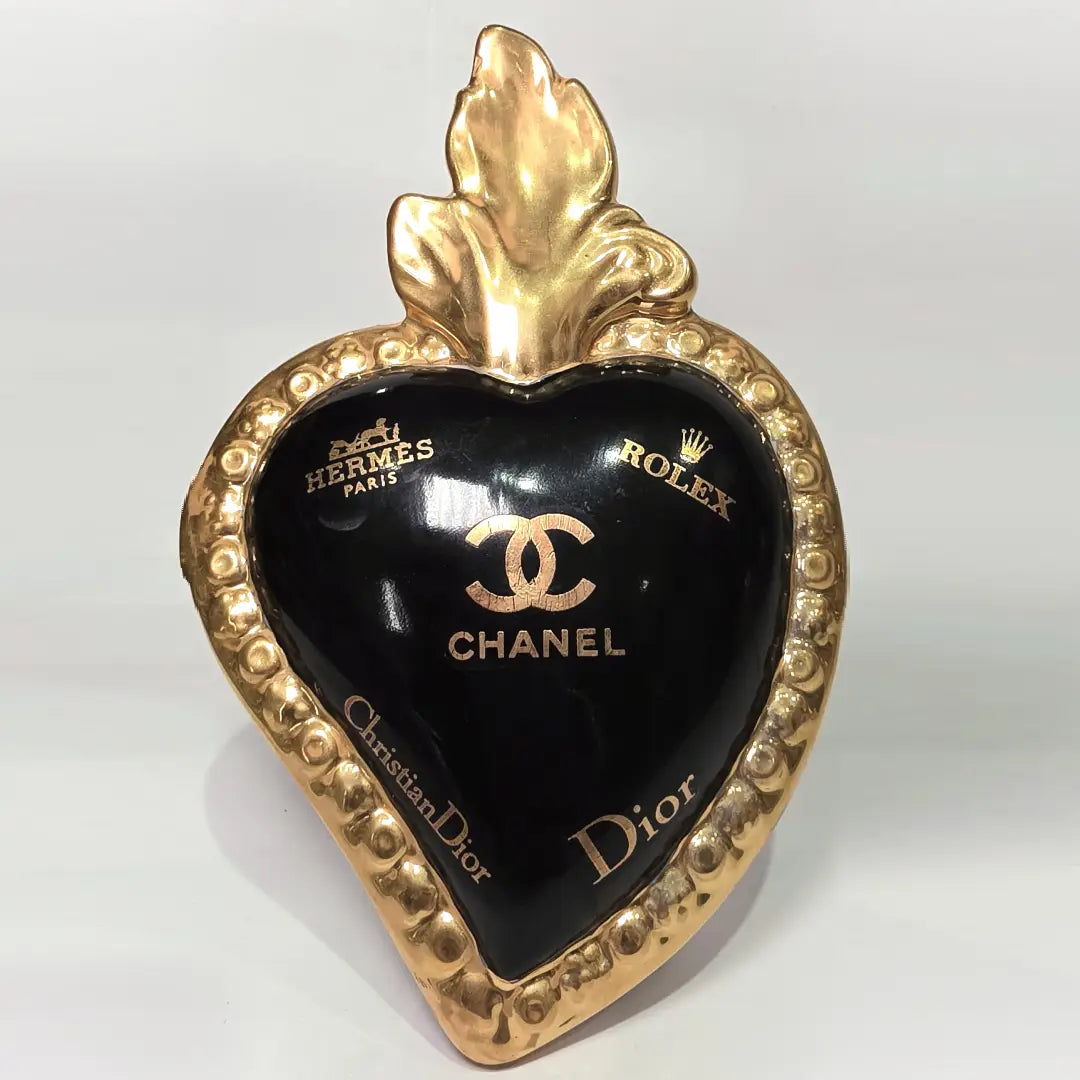 Scultura a forma di Cuore Sacro in ceramica nera lucida, impreziosita da una ricca cornice dorata lavorata a sbalzo e decorata con loghi fashion iconici in oro. Questo esclusivo oggetto d'arredo in stile pop art presenta una fiamma superiore dorata e una base d'appoggio integrata, perfetto per aggiungere un dettaglio di lusso moderno agli ambienti interni.