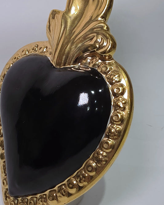 Scultura a forma di Cuore Sacro in ceramica nera lucida, incorniciata da un elegante bordo dorato con decorazioni a rilievo e completata da una fiamma superiore in oro. Questo oggetto di design artigianale con base d'appoggio è un complemento d'arredo raffinato, ideale per arricchire ambienti moderni con un tocco di arte sacra contemporanea.