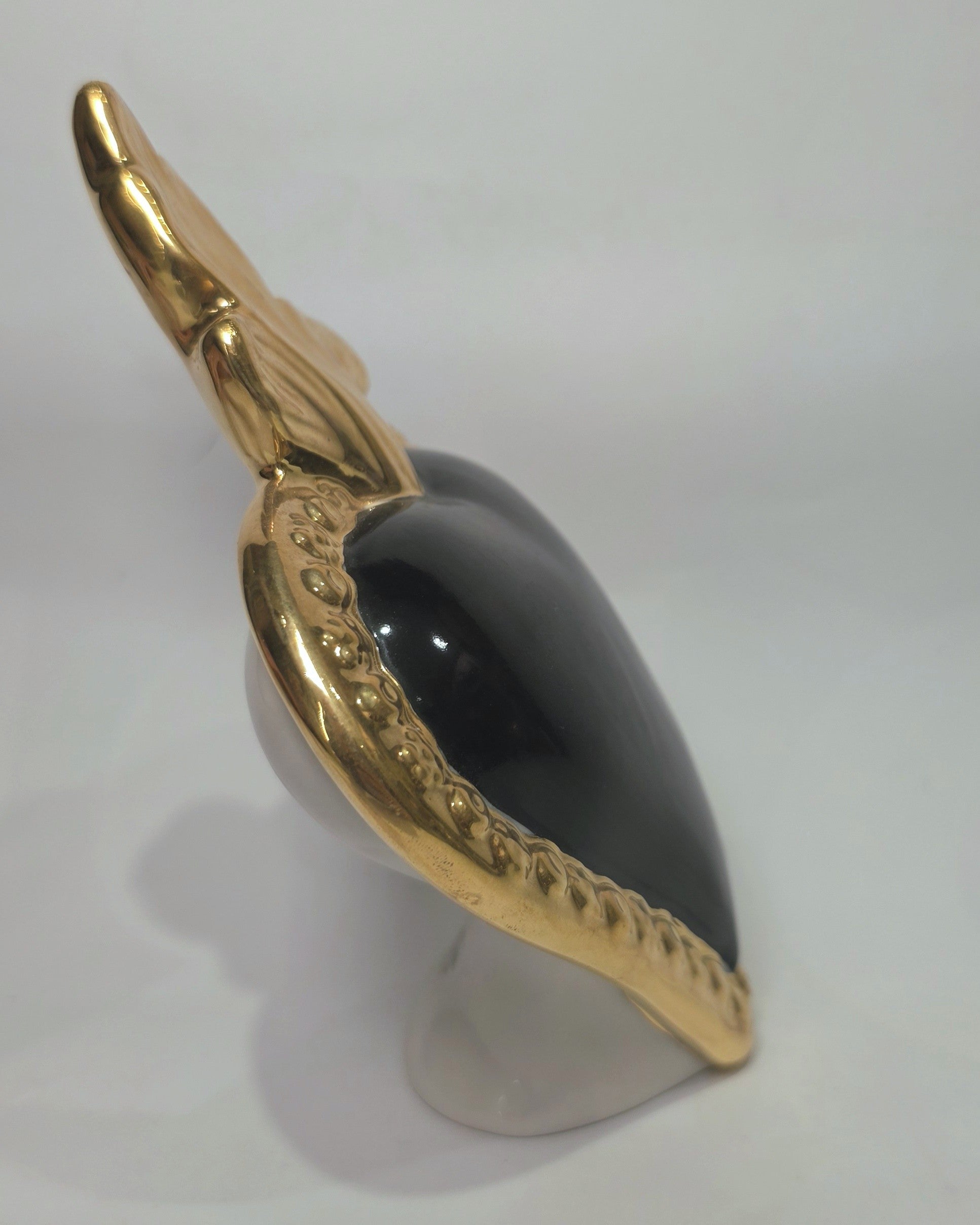 Scultura a forma di Cuore Sacro in ceramica nera lucida, incorniciata da un elegante bordo dorato con decorazioni a rilievo e completata da una fiamma superiore in oro. Questo oggetto di design artigianale con base d'appoggio è un complemento d'arredo raffinato, ideale per arricchire ambienti moderni con un tocco di arte sacra contemporanea.