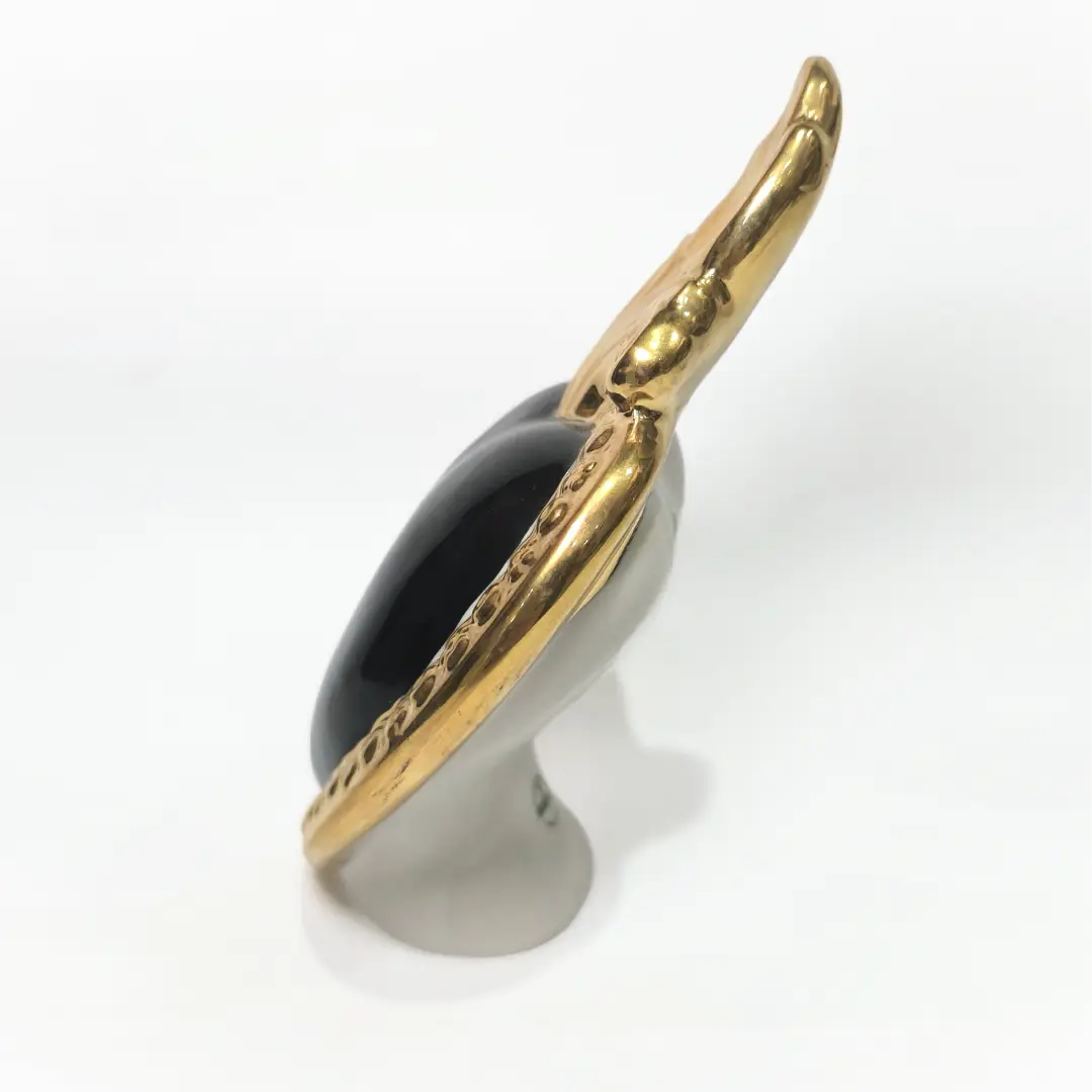 Scultura a forma di Cuore Sacro in ceramica nera lucida, incorniciata da un elegante bordo dorato con decorazioni a rilievo e completata da una fiamma superiore in oro. Questo oggetto di design artigianale con base d'appoggio è un complemento d'arredo raffinato, ideale per arricchire ambienti moderni con un tocco di arte sacra contemporanea.