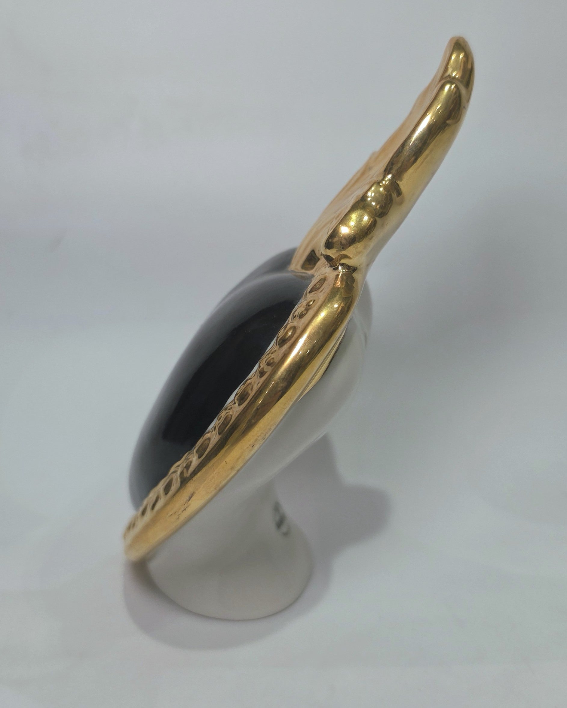 Scultura a forma di Cuore Sacro in ceramica nera lucida, incorniciata da un elegante bordo dorato con decorazioni a rilievo e completata da una fiamma superiore in oro. Questo oggetto di design artigianale con base d'appoggio è un complemento d'arredo raffinato, ideale per arricchire ambienti moderni con un tocco di arte sacra contemporanea.
