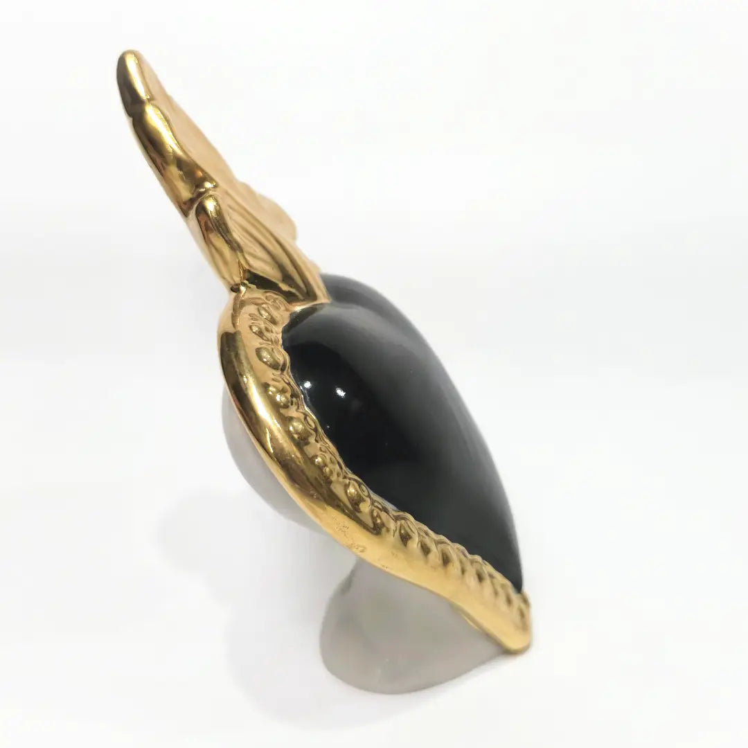 Scultura a forma di Cuore Sacro in ceramica nera lucida, incorniciata da un elegante bordo dorato con decorazioni a rilievo e completata da una fiamma superiore in oro. Questo oggetto di design artigianale con base d'appoggio è un complemento d'arredo raffinato, ideale per arricchire ambienti moderni con un tocco di arte sacra contemporanea.